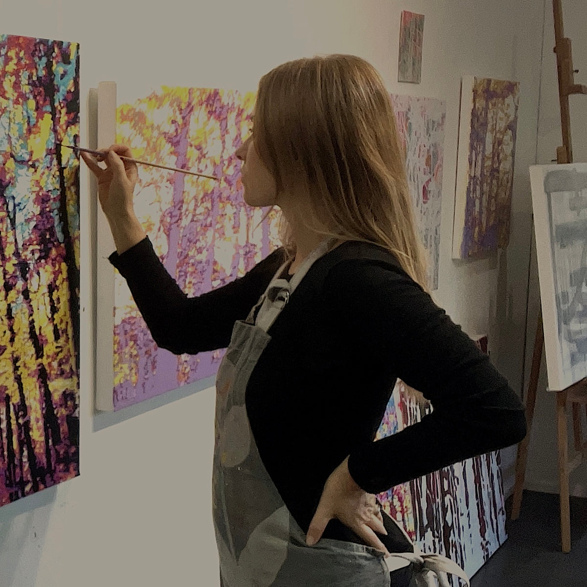 Artist Alex DiMercurio painting in a black long-sleeve shirt and gray painter's overalls stands in profile, holding a paintbrush and painting an abstract tree artwork on a canvas in her art studio.